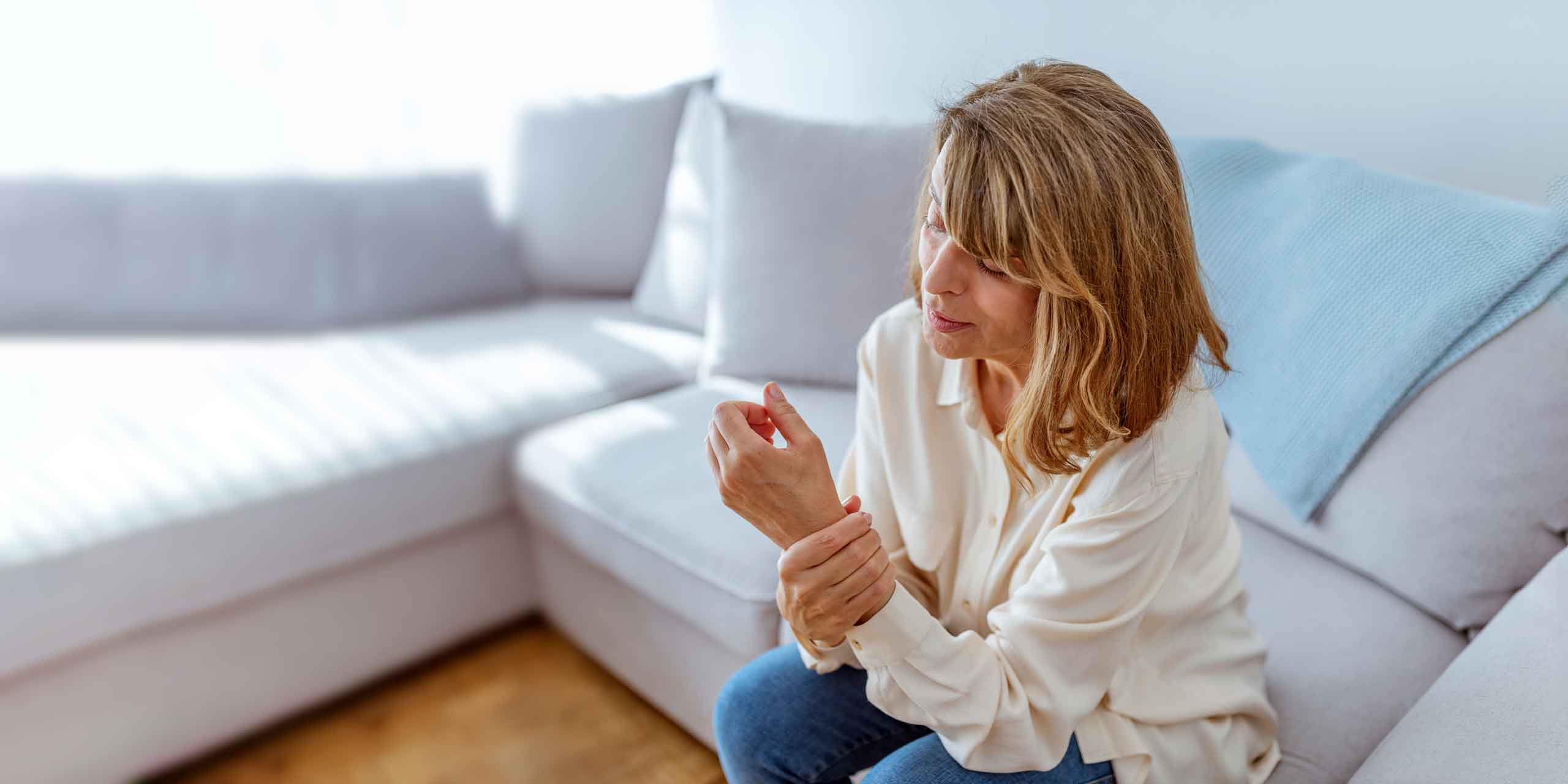Eine Frau in weißer Bluse und Jeans sitzt in einem hell erleuchteten Wohnzimmer auf einer Couch und blickt mit besorgtem Gesichtsausdruck auf ihre Hand.