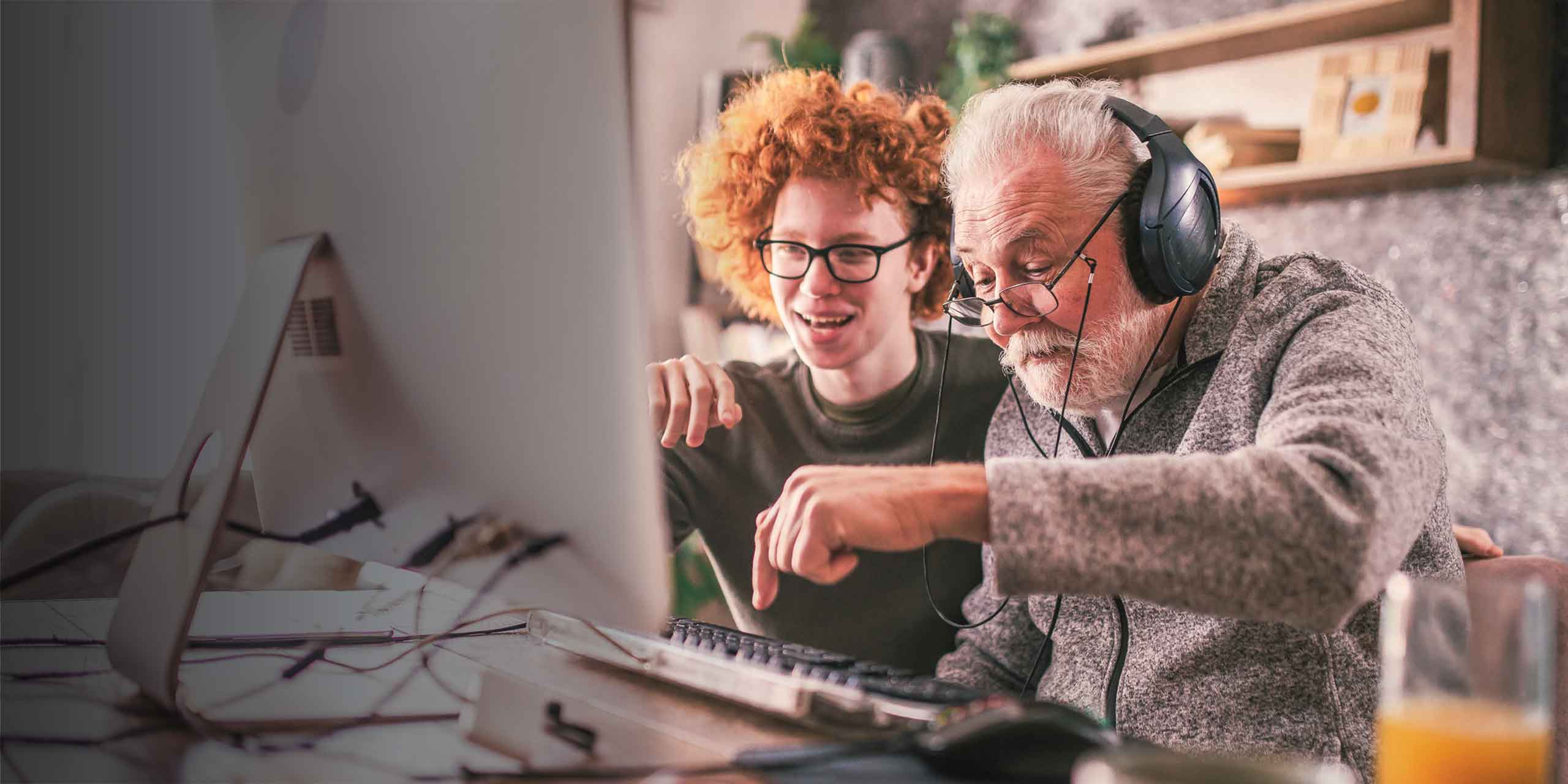 Ein älterer Herr mit Kopfhörern und ein junger Erwachsener mit lockigem rotem Haar lächeln und zeigen auf einen Computerbildschirm. Sie sind in einem gemütlichen Raum gemeinsam einer Aktivität nachgegangen.