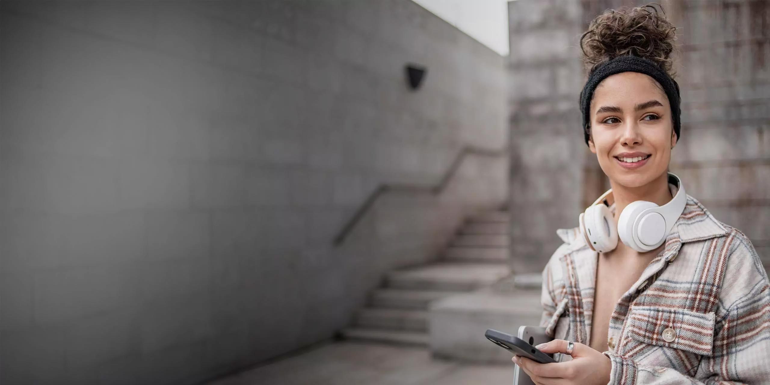 Eine junge Frau mit Stirnband und Kopfhörern um den Hals lächelt, während sie ein Smartphone in der Hand hält und auf einer Treppe in einer städtischen Umgebung sitzt.