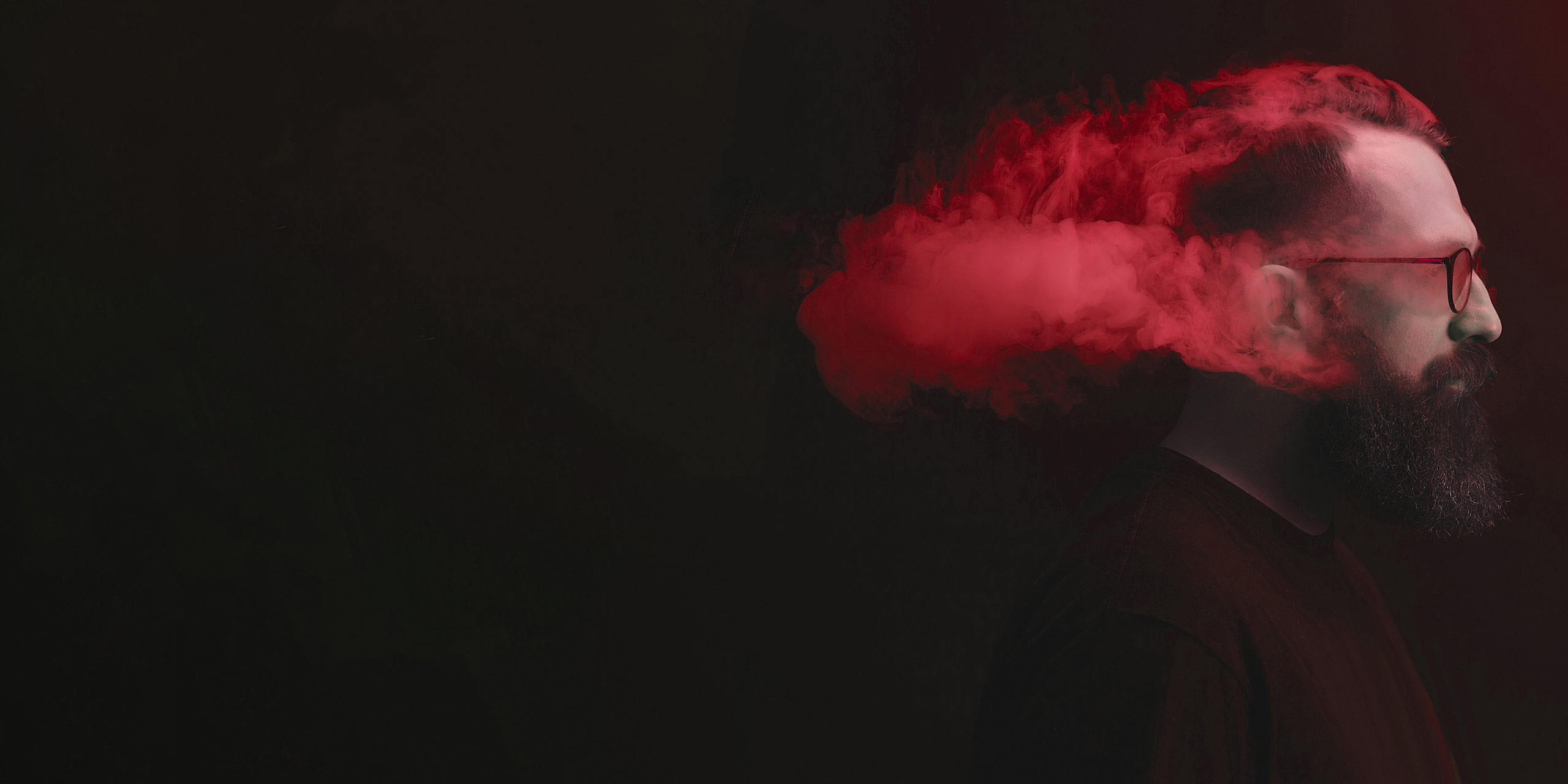 Bärtiger Mann mit Brille im Profil mit rotem Rauch um den Kopf vor schwarzem Hintergrund