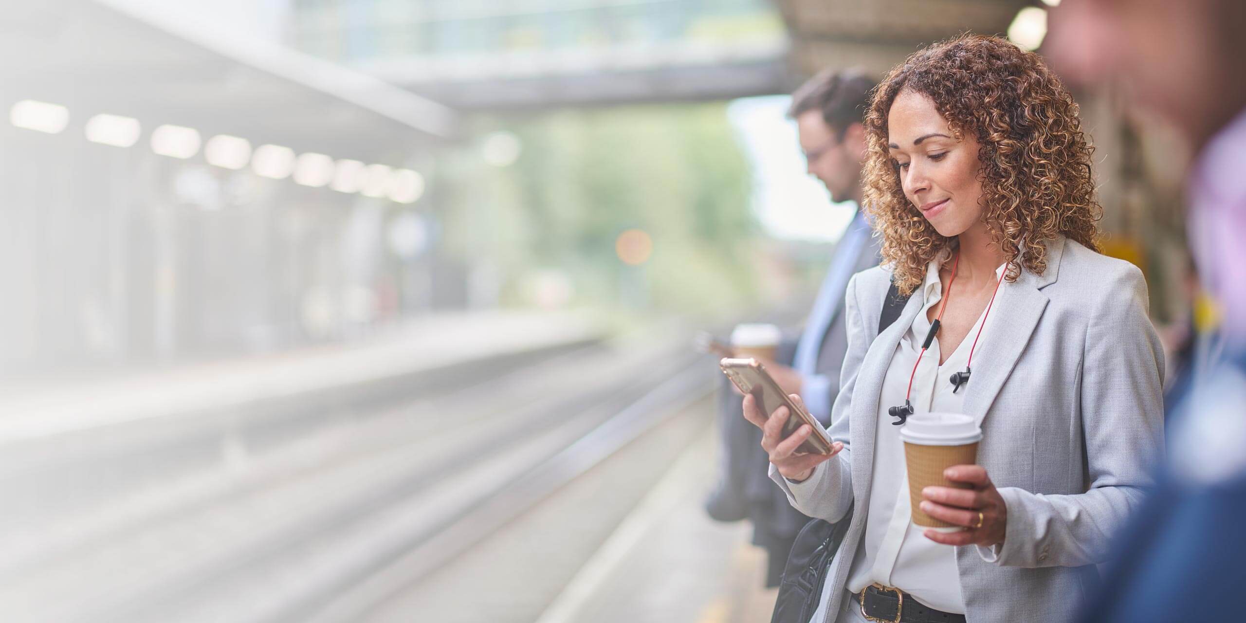 Eine Frau steht auf einem Bahnsteig, hält eine Kaffeetasse in der Hand und schaut auf ihr Smartphone. Sie trägt einen hellgrauen Blazer und hat Ohrstöpsel im Ohr. Im Hintergrund sind weitere Passagiere zu sehen, die ebenfalls auf den Zug warten. Die Gleise und der Bahnsteig sind zu sehen.