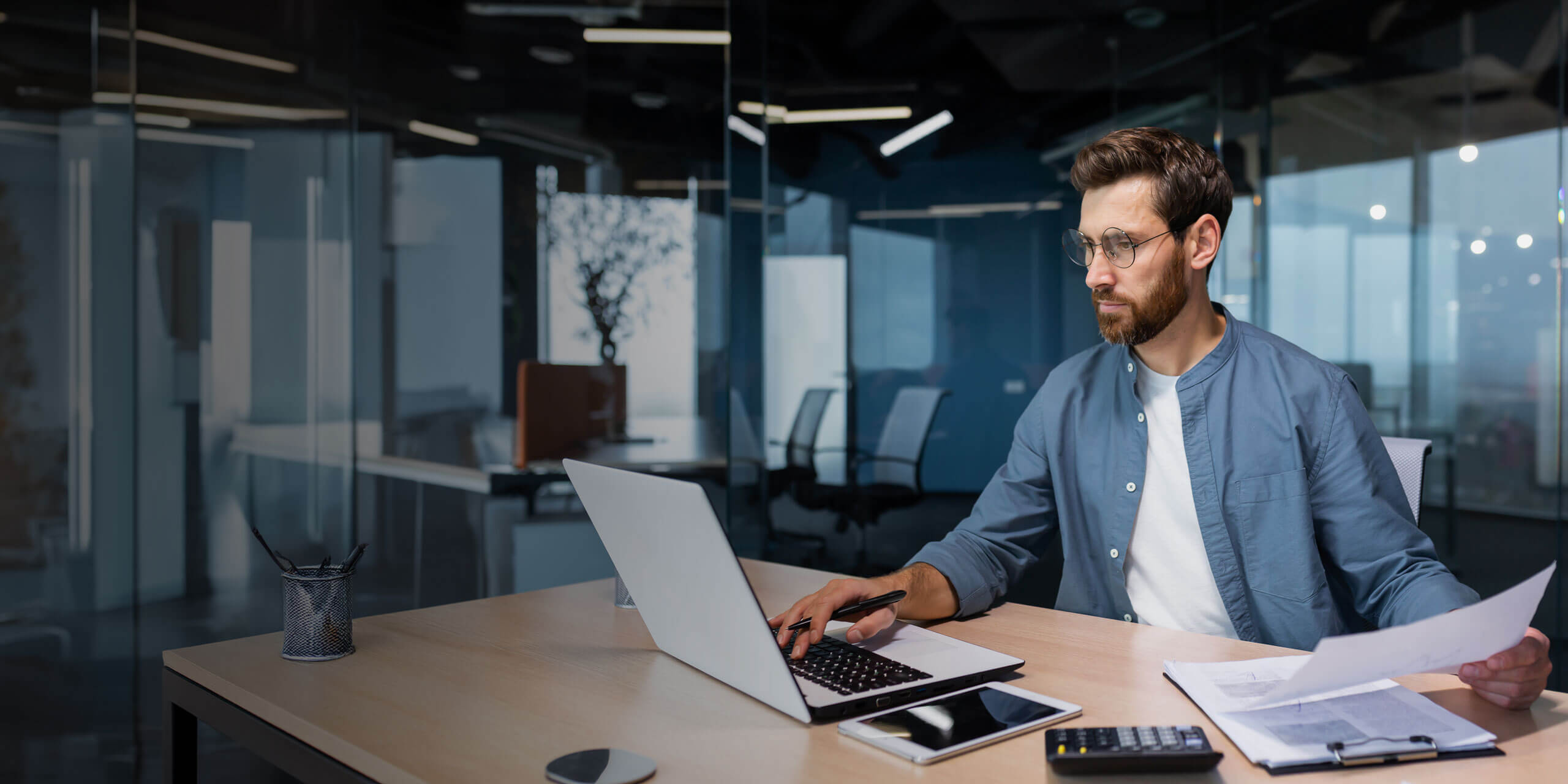 Ein Mann mit Brille und Bart arbeitet an einem Schreibtisch in einem Büro. Er benutzt einen Laptop und hält ein Blatt Papier in der Hand, während ein Taschenrechner, ein Tablet, ein Smartphone und ein Notizblock auf dem Schreibtisch liegen. Das Büro hat Glaswände und ist schwach beleuchtet.