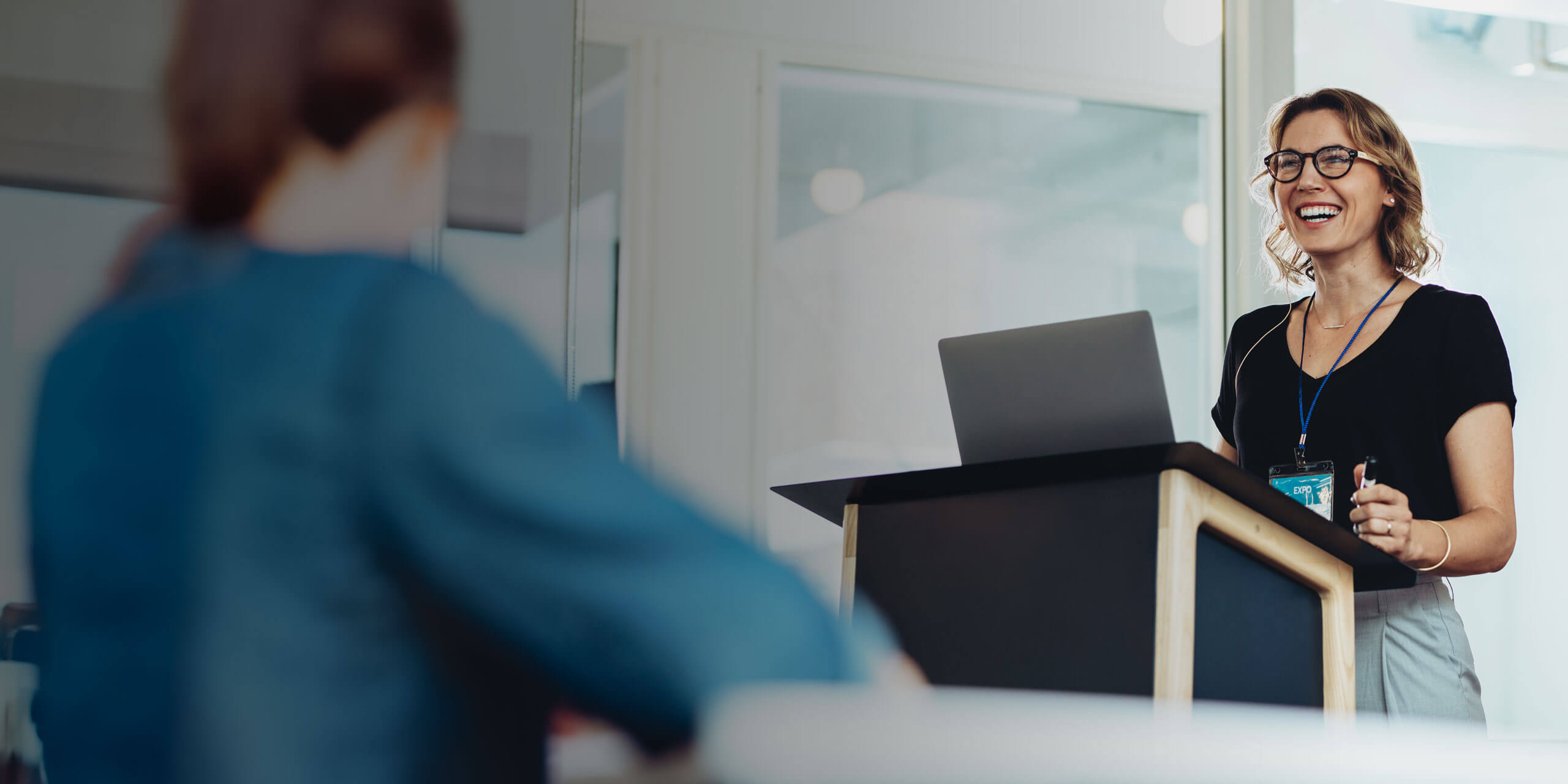 Eine Frau steht lächelnd mit einem Laptop hinter einem Rednerpult und spricht zu einem sitzenden Publikum in einem hellen, modernen Büroraum. Sie trägt eine Brille und ein schwarzes Hemd und schafft so eine professionelle und ansprechende Atmosphäre.